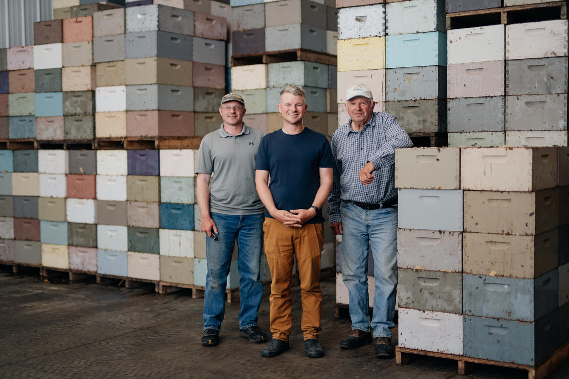 Three men stand in front of palettes of honey
