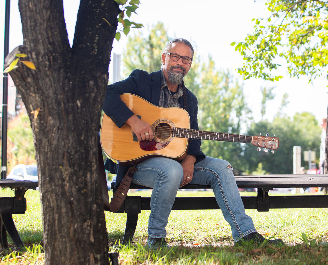 Craig Ginn sits on a picnic table in a sunny park with his guitar