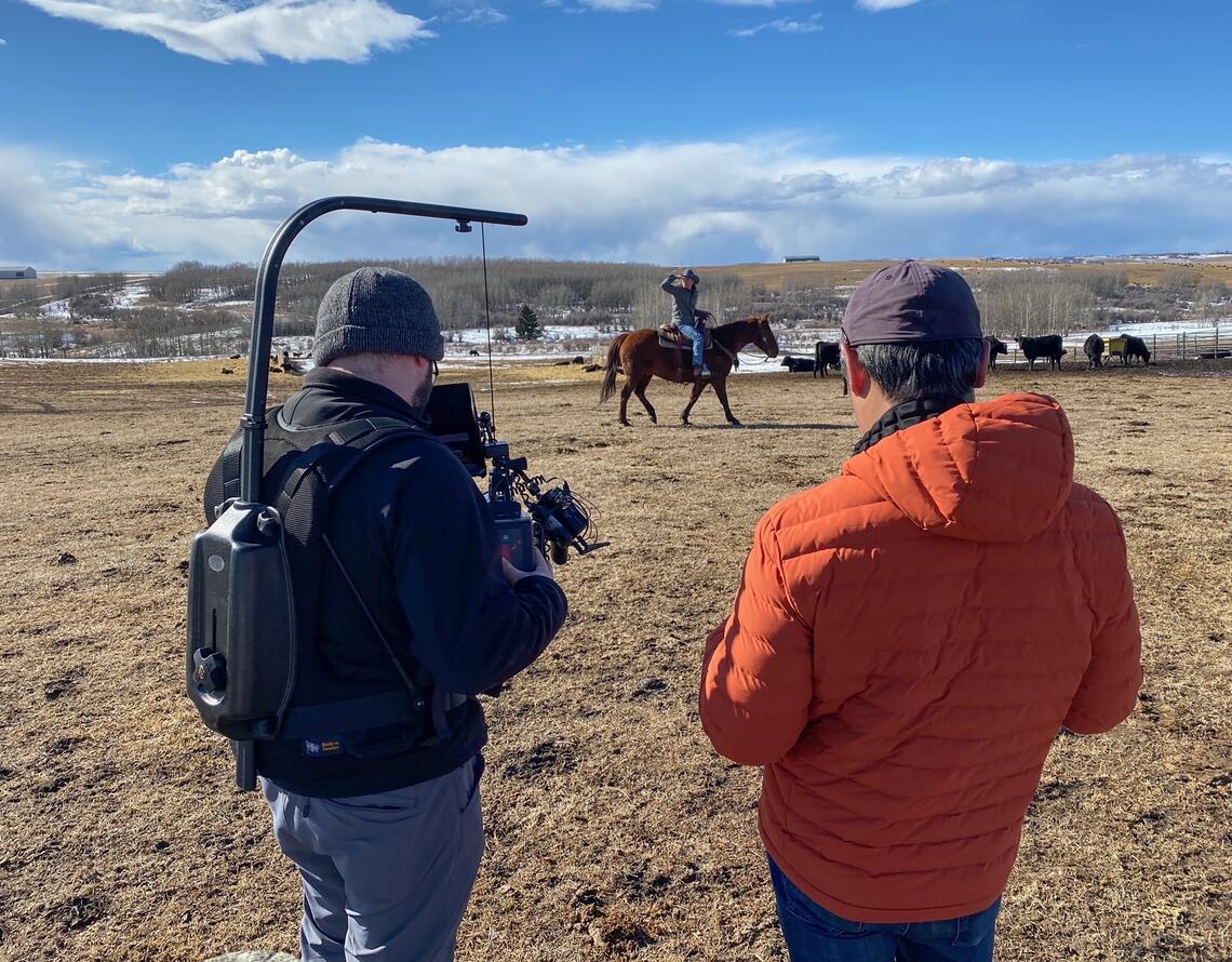 A two-person crew films a man riding a horse outdoors on a cattle ranch