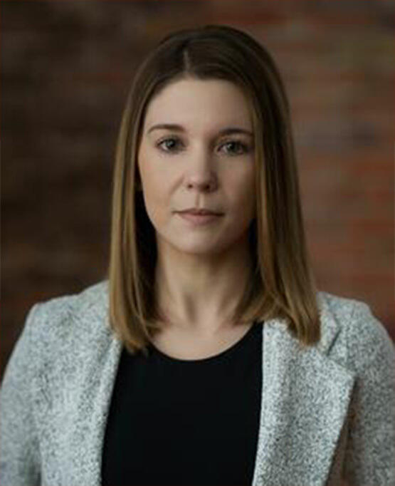 A woman stands facing the camera with a brick wall behind her