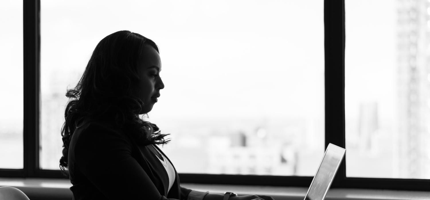 Woman sitting in front of a laptop in front of windows overlooking a city.