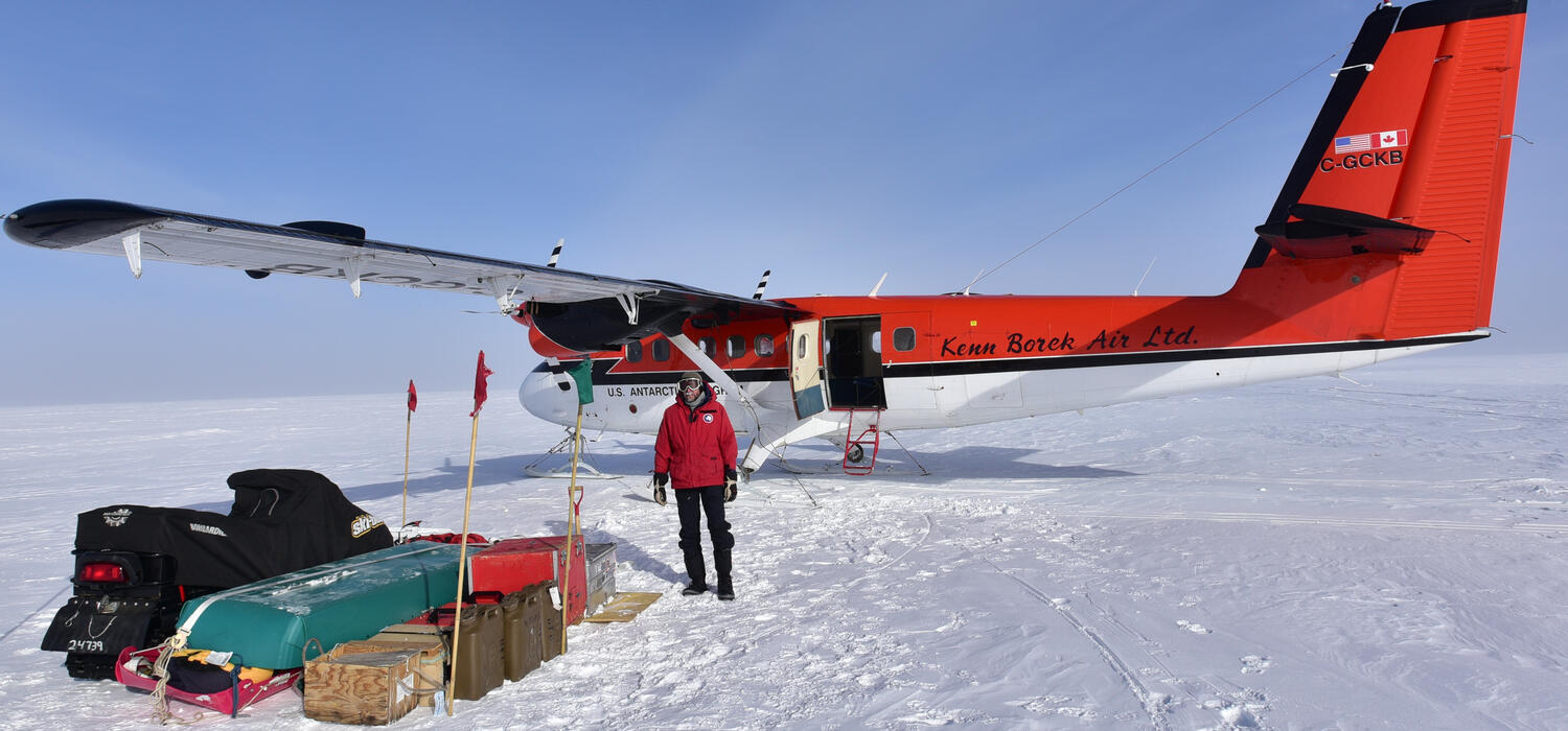 A man in a red jacket stands in front of a red plane with luggage stacked beside him on a snowy landscape