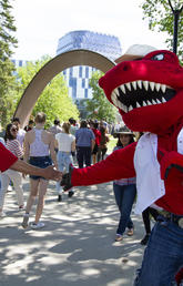 UCalgary's Mascot Rex high-fives a man in a jean jacket as he waits in line for the President's Stampede BBQ on July 12. The two are pictured in the quad on main campus with the TFDL in the background. A woman smiles in the background as she watches the high-five.