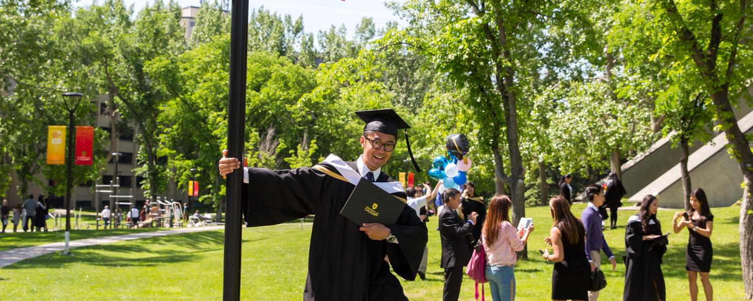 A graduate holding his degree in one hand and posing with a lamp post with the other hand 