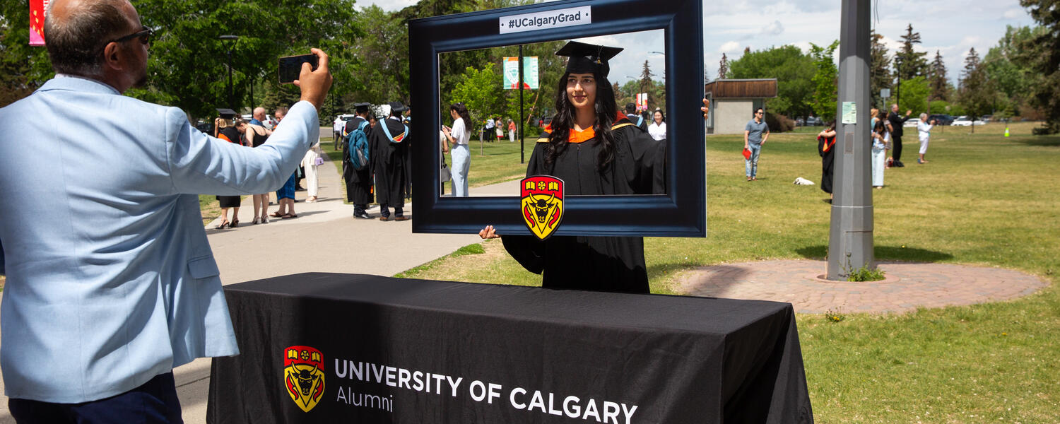 A grad holds up an alumni frame while someone takes a photo of them outdoors at UCalgary.