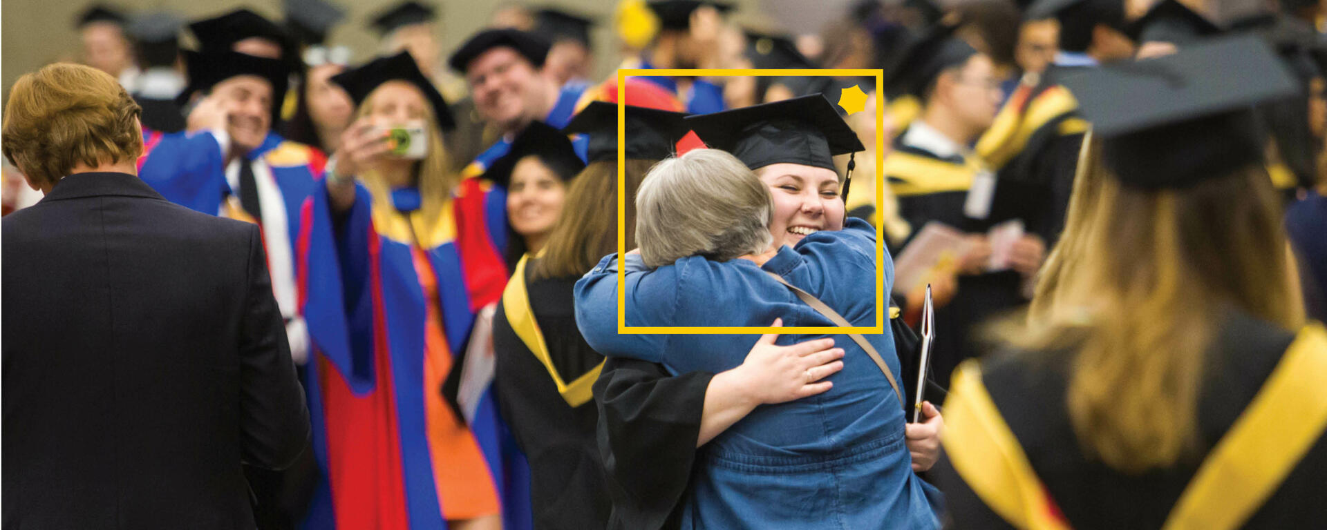 A photo of two people embracing during convocation with a yellow Alumni square over top.