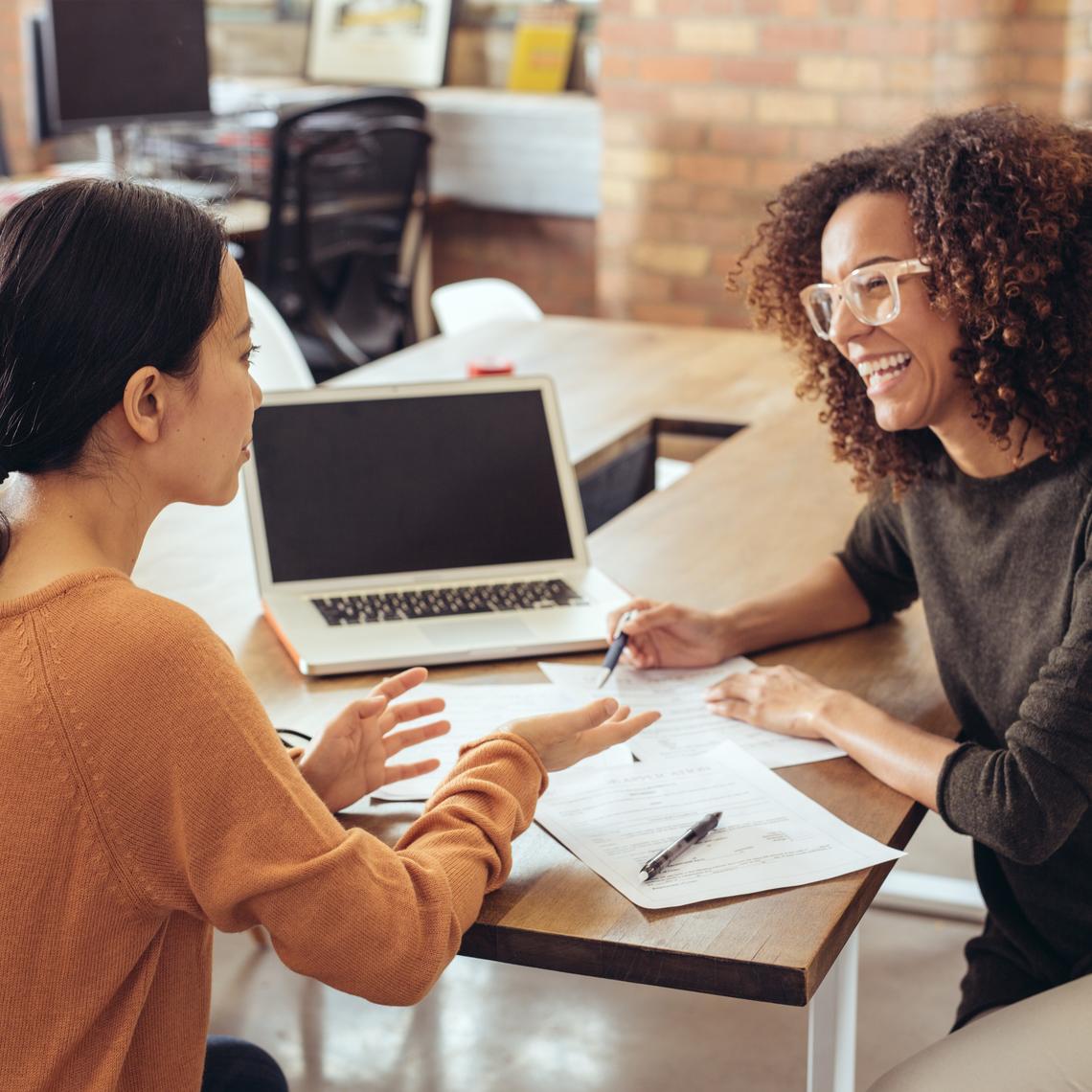 Two women talking with each other with laptop and papers