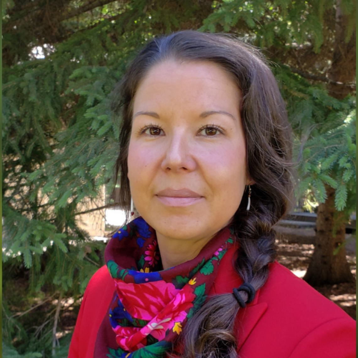 A photo of Kori Czuy wearing a red jacket in front of a green tree. 