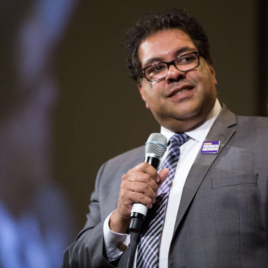 Mayor Nenshi wearing a grey suit and holding a microphone on stage at an event.