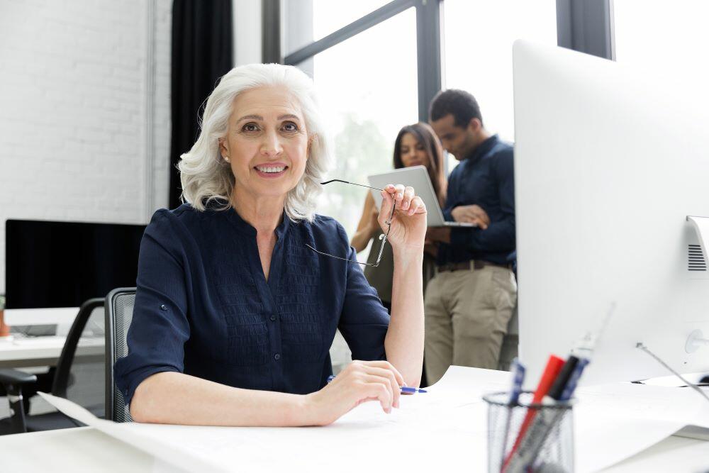 A woman with grey hair sits at a computer desk with other coworkers in the background