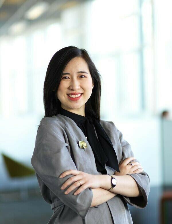 A photo of Fay wearing a grey blazer and black shirt in front of large windows.