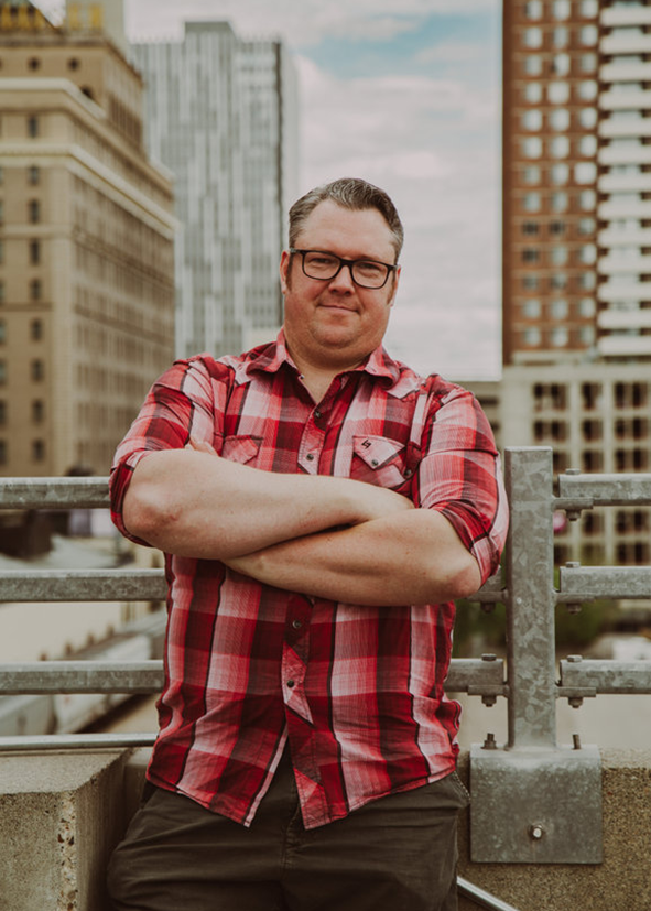 A photo of Stephen in a red plaid shirt on a rooftop with buildings in the background