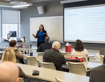 Alumni counsellor Lorelei stands at the front of a room speaking to an audience. On the screen a slide that says "what is burnout" is visible.